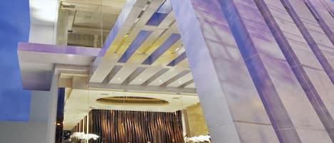 Un hôtel moderne, avec un grand nombre d’étages, dont la façade est remarquable grâce à son parement en verre, un hall d’accueil doté d’un comptoir jaune et un escalier bien visible menant aux étages supérieurs.