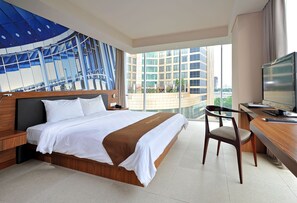 Habitación de hotel moderna con una cama grande, un escritorio con computadora y vistas a la ciudad a través de amplias ventanas.