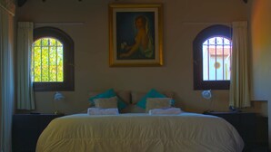 A bedroom with a large bed, two bedside tables, a painting above the bed, and two windows with curtains. - Susana Just B&B (Florida)