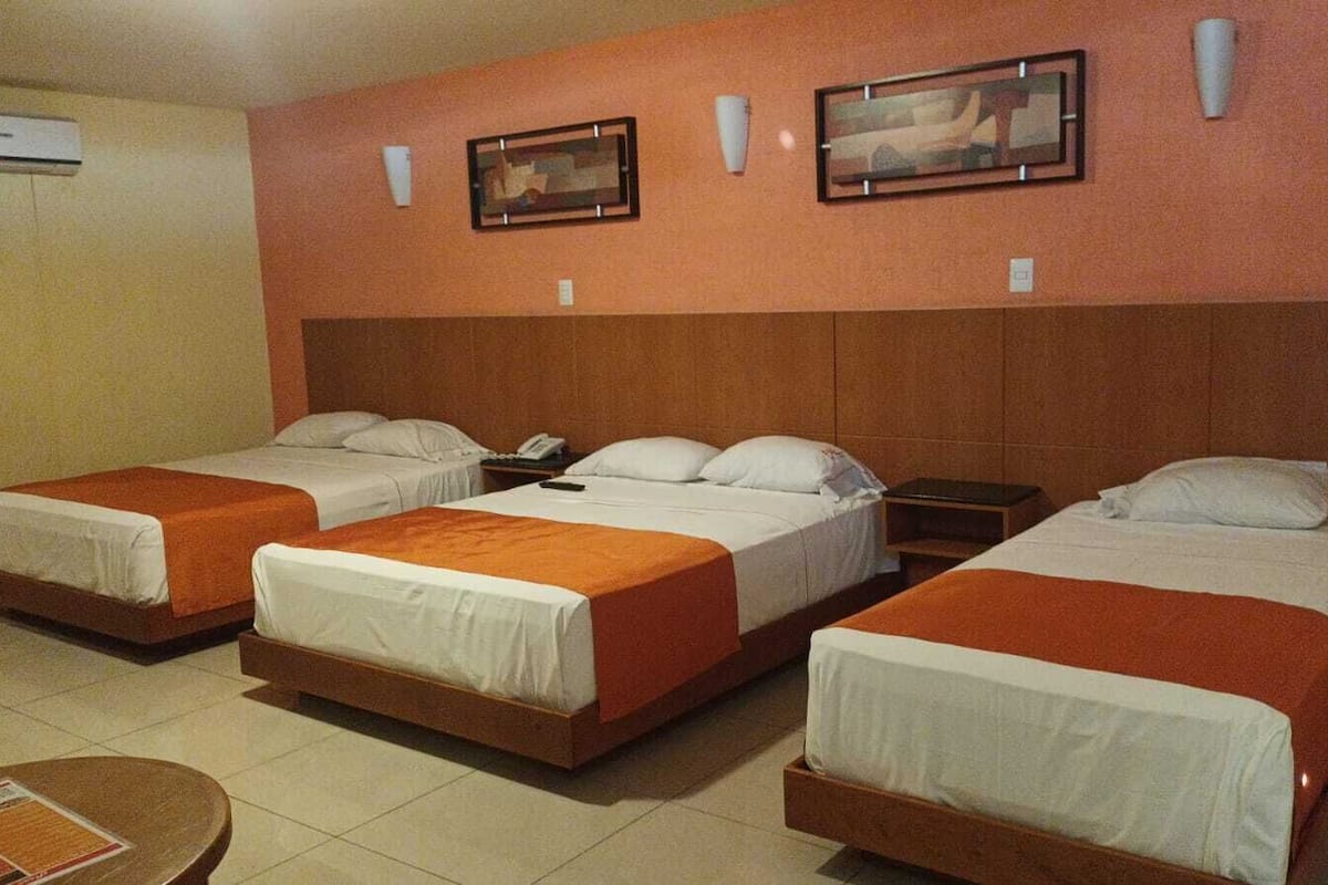Ein Hotelzimmer mit drei Einzelbetten, einem kleinen Tisch und einer Wand-Klimaanlage.