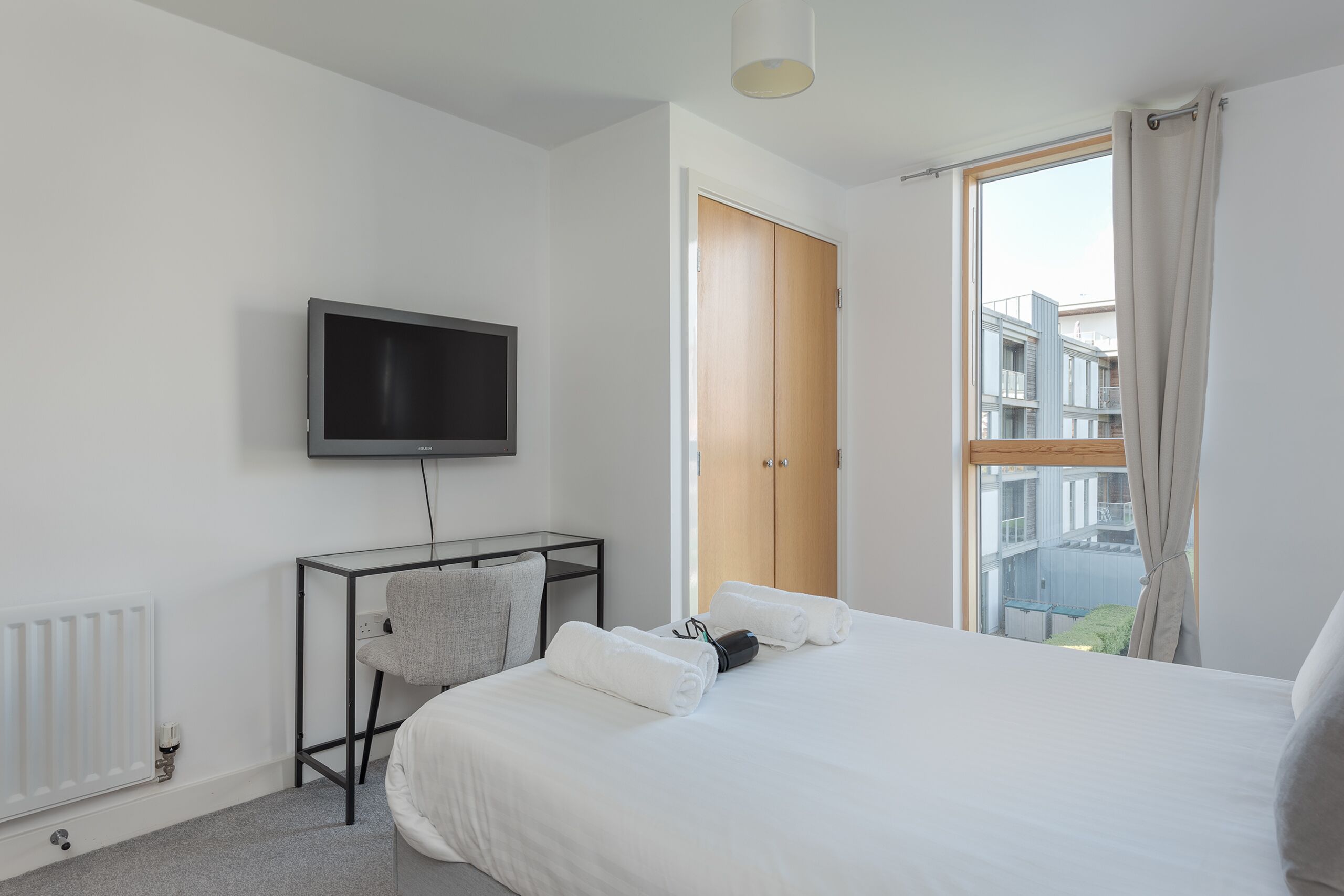 Une chambre d’hôtel moderne équipée d’un lit, d’un téléviseur fixé au mur, d’un bureau avec une chaise et d’une grande fenêtre avec des rideaux.