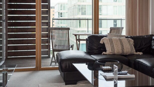 Un salon moderne avec un canapé en cuir noir, une table basse en verre et une vue sur le paysage urbain, visible à travers de grandes fenêtres.
