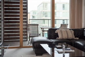 Een moderne woonkamer met een zwarte leren bank, een glazen salontafel en uitzicht op een stadsgezicht door grote ramen.