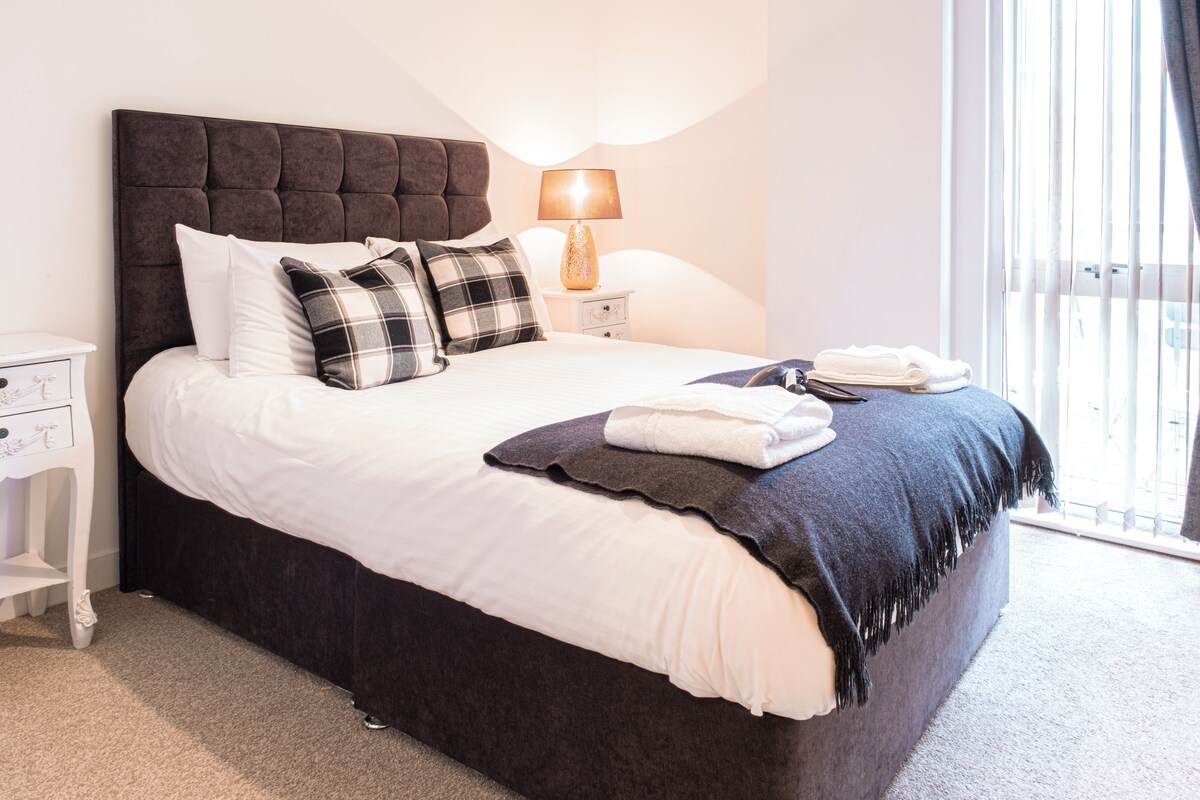 Una cama bien tendida con cabecera oscura, ropa de cama blanca y varios cojines decorativos. Hay una mesita de noche con una lámpara y una toalla doblada. La habitación tiene un ventanal con persianas.