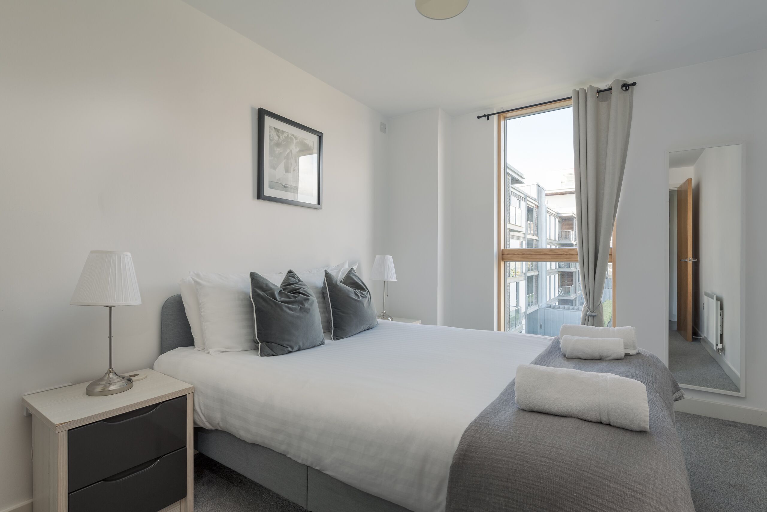 Une chambre d’hôtel moderne avec un grand lit, deux tables de chevet, une fenêtre avec des rideaux, un tableau encadré au mur et une vue sur la ville.