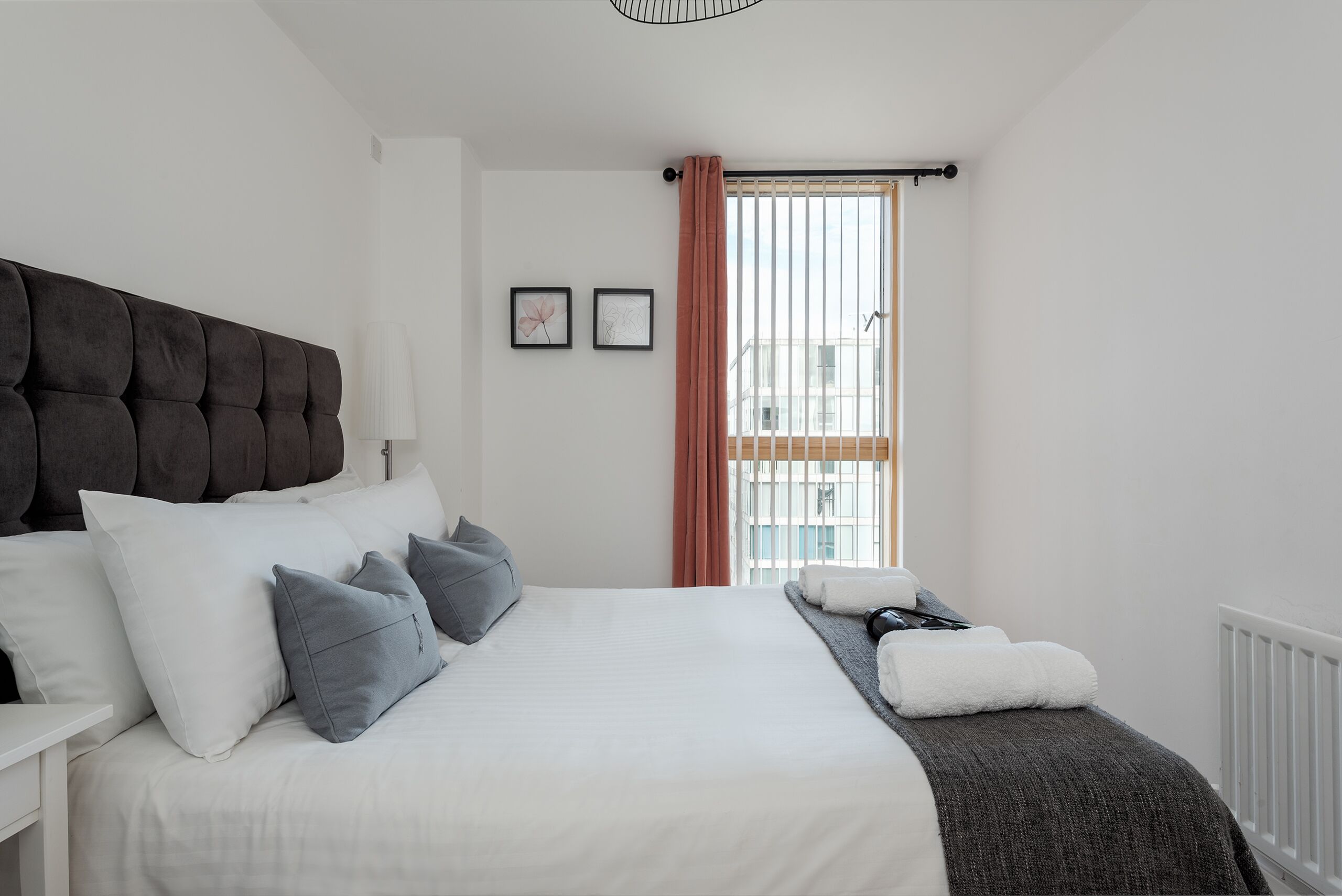 Ein ordentlich gemachtes Bett mit Kissen, ein Nachttisch, ein Fenster mit Vorhängen und eine Heizung.