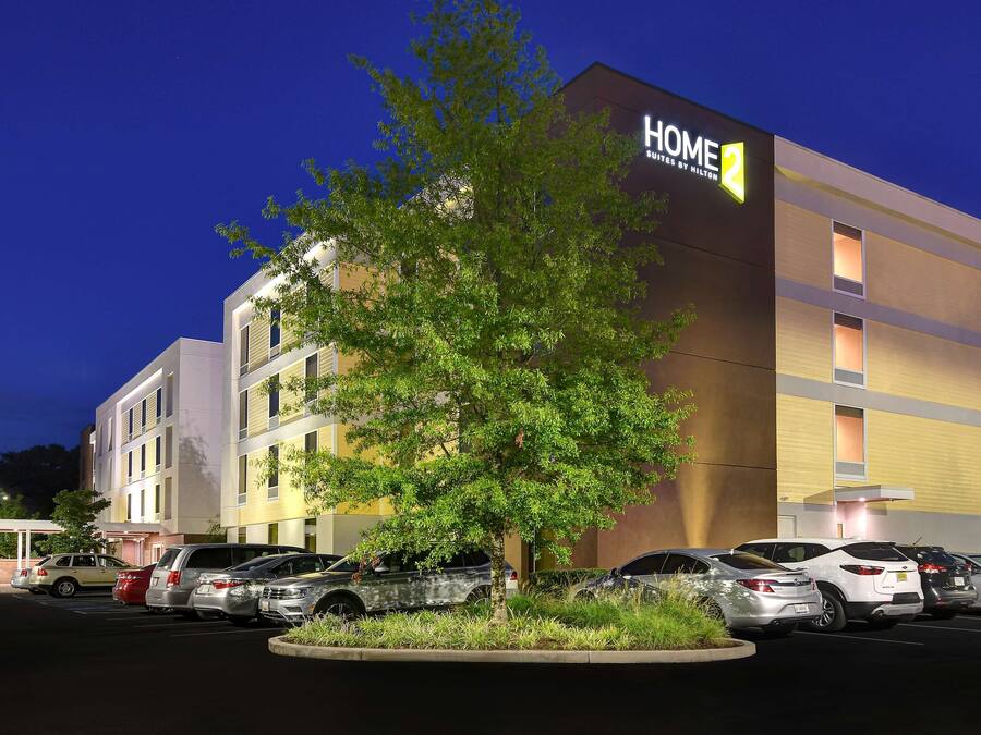 Un bâtiment moderne abritant un hôtel, avec une enseigne indiquant « HOME 2 » et un parking où sont stationnées plusieurs voitures.