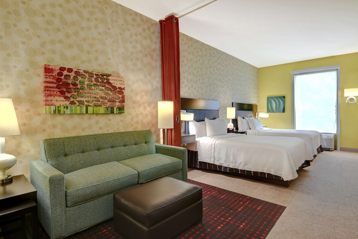 A hotel room with a green sofa, a bed with white linens, a red curtain, and a patterned carpet.