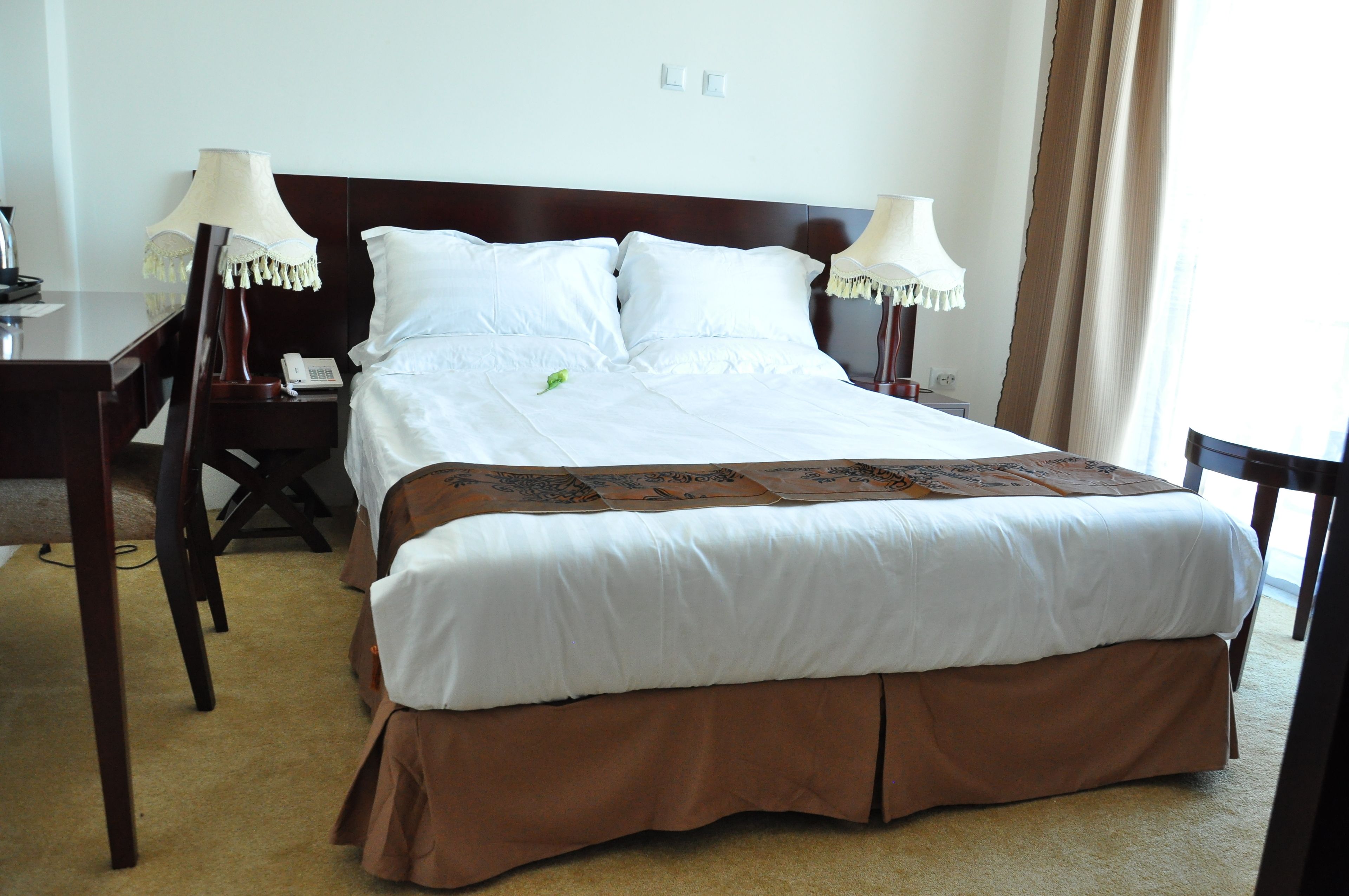 A neatly made bed with white linens and a brown skirt, two bedside lamps, a telephone, and a small table with a lamp.
