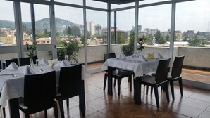 A restaurant with tables set for dining, large windows offering a city view, and potted plants on the tables. - Ag Palace Hotel (Addis Ababa)