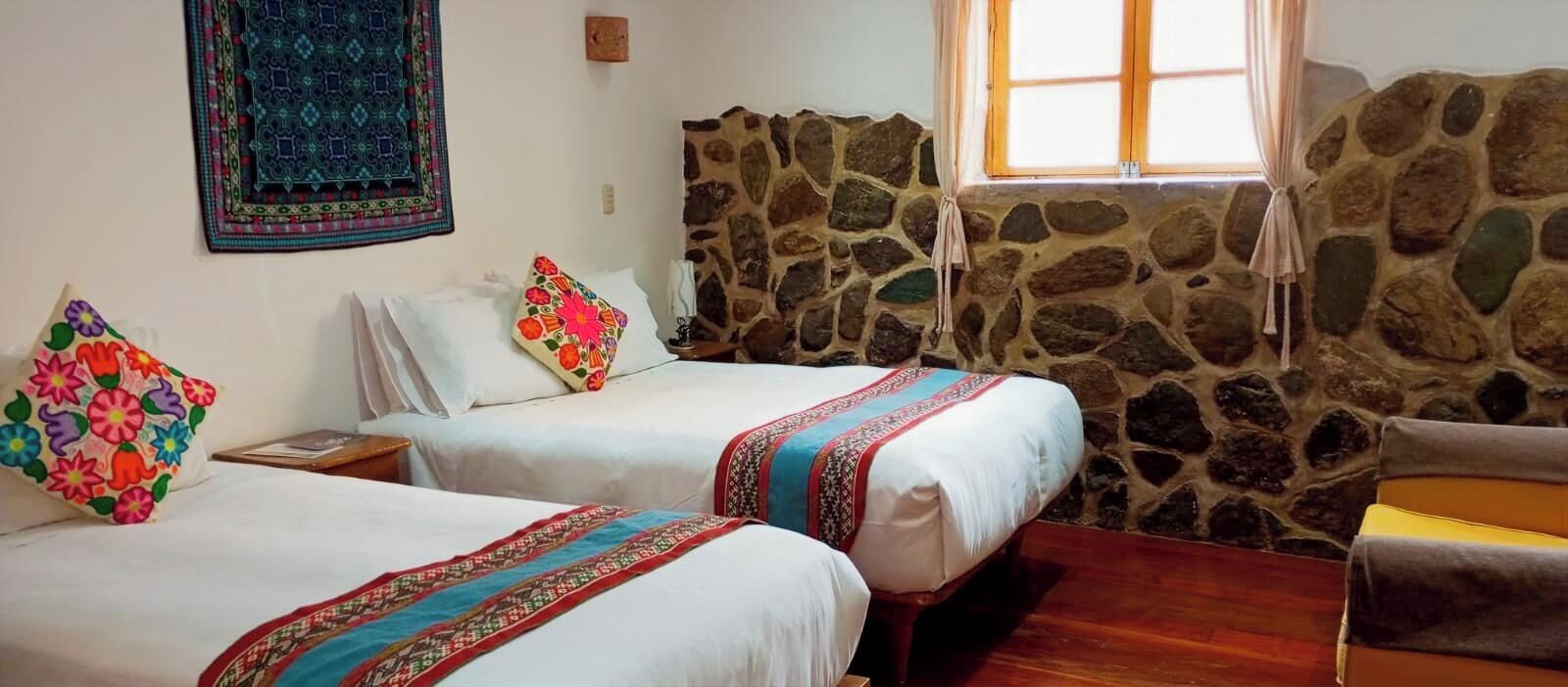 Una habitación con dos camas, una pared de piedra, una ventana con cortinas y una mesita pequeña.