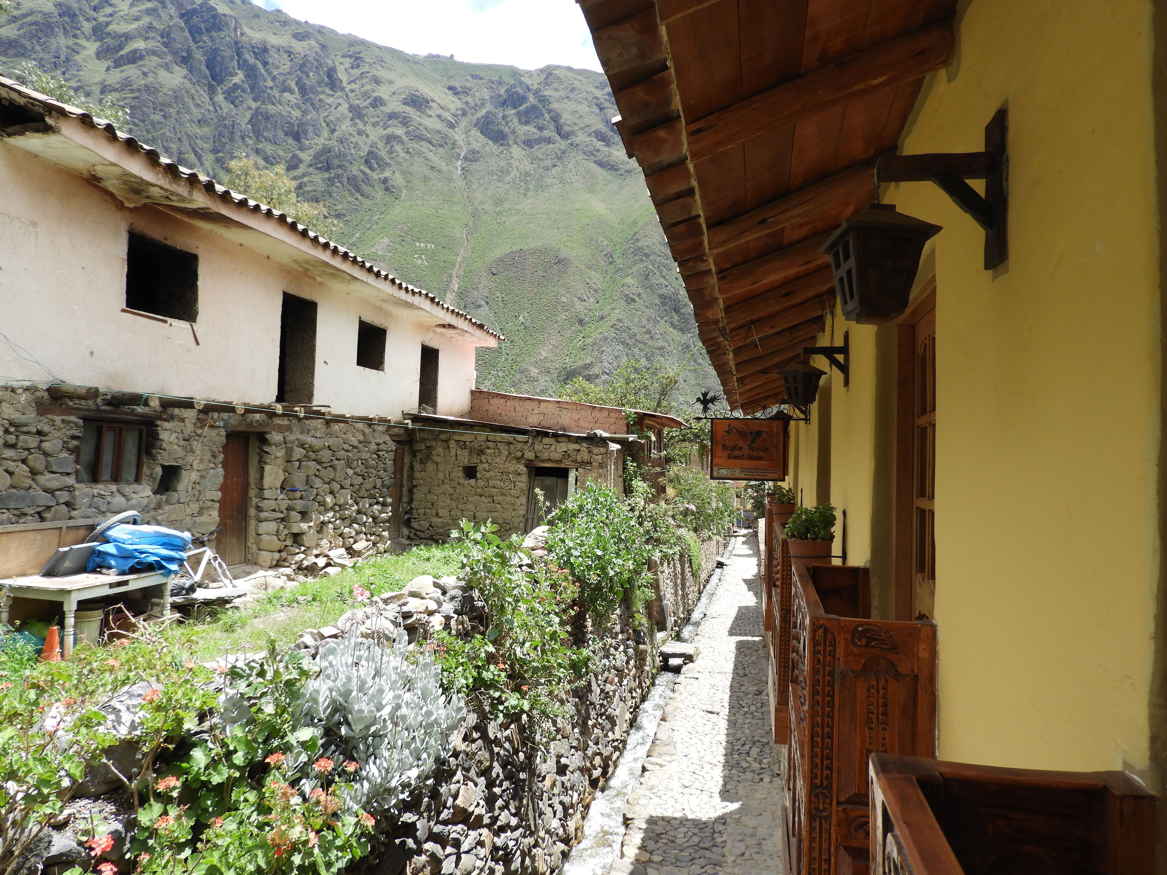 A narrow cobblestone street with stone buildings, a wooden fence, and a mountain backdrop.