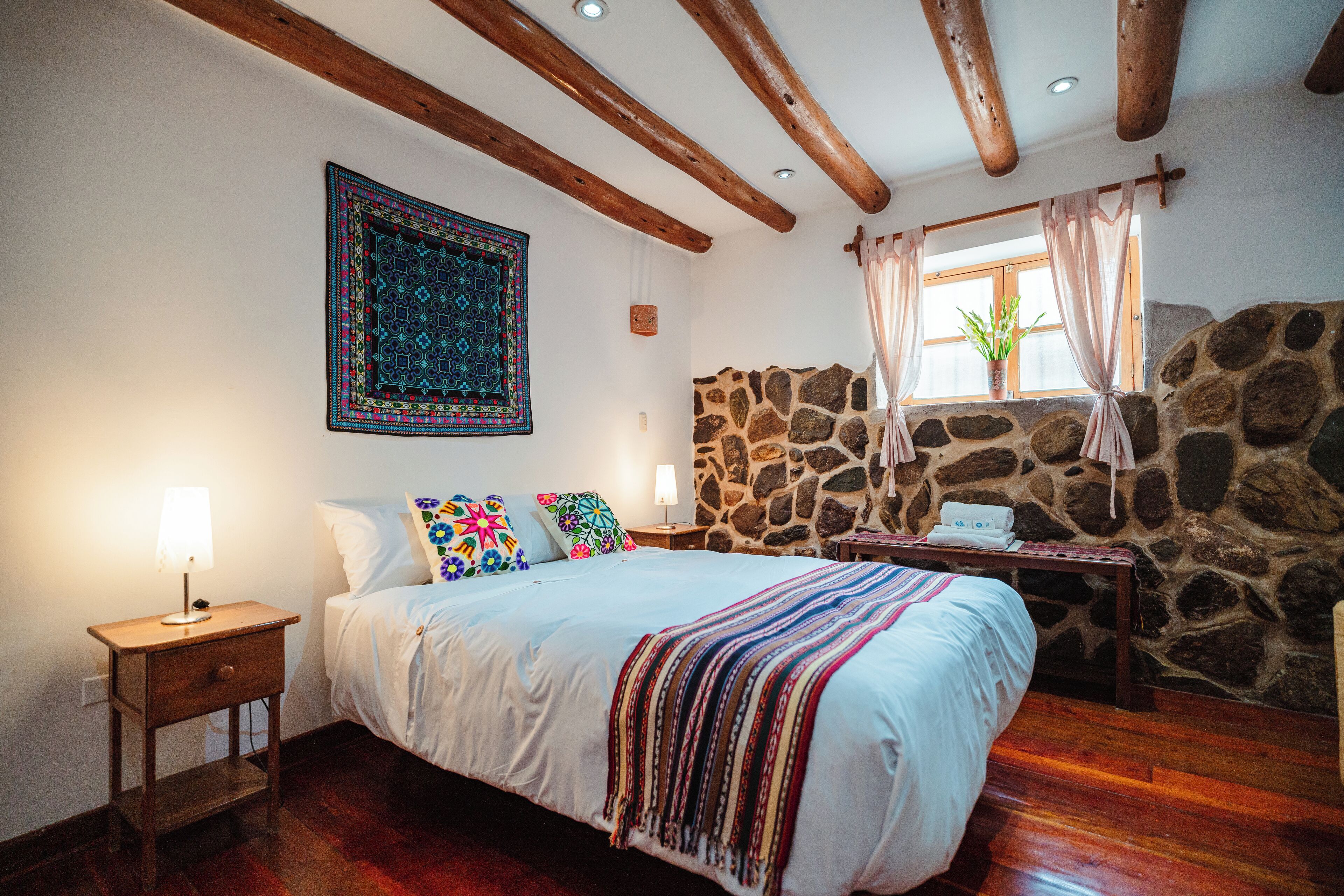 A bedroom with a large bed, a nightstand, a stone wall, and a window with curtains.