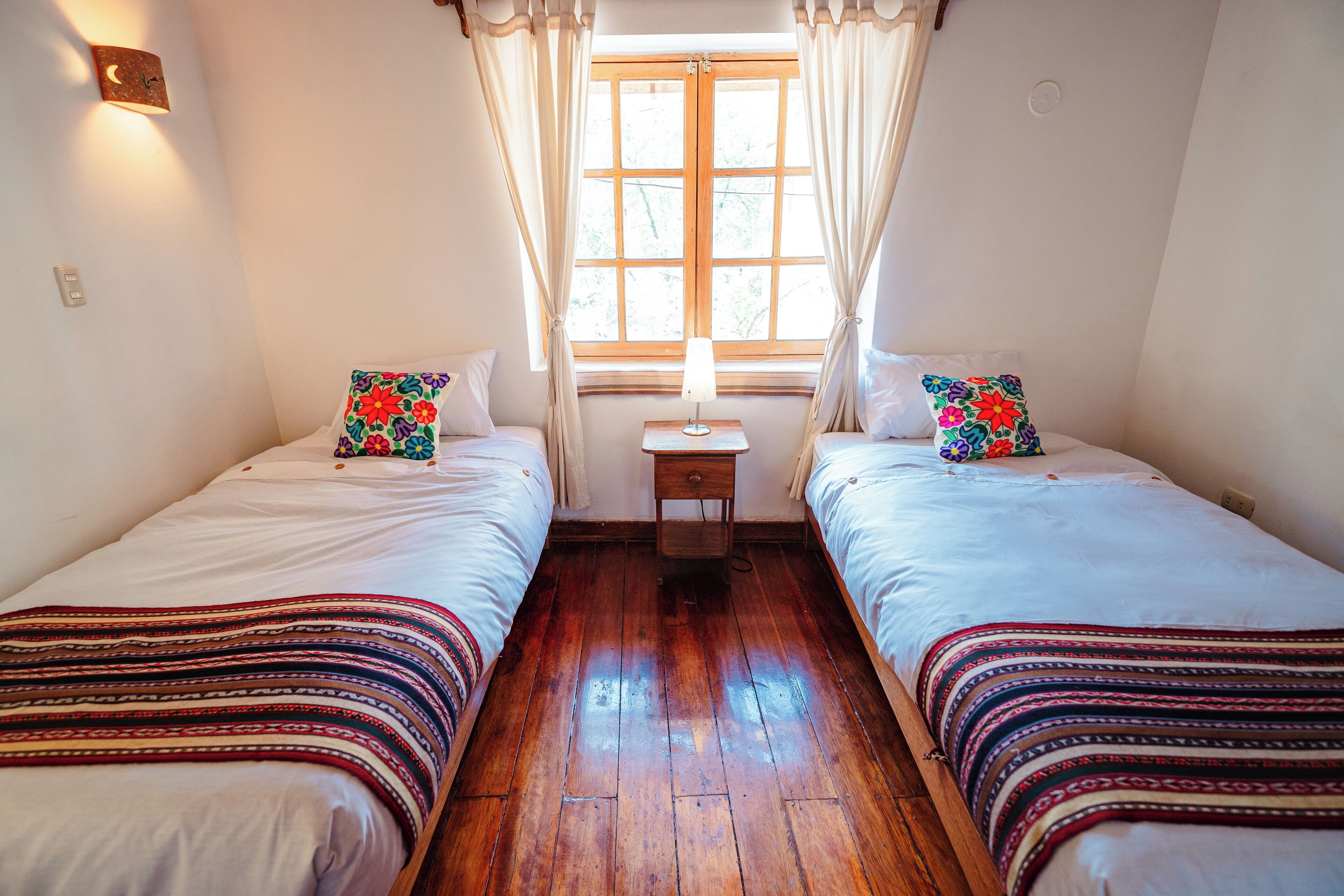 Una habitación con dos camas, cada una con un cojín de colores, una mesita de noche pequeña con una lámpara y una ventana con cortinas.
