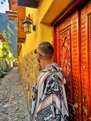 A person standing in front of a wooden door with intricate carvings, a stone wall, and a cobblestone path. - B&B Picaflor Tambo (Ollantaytambo)