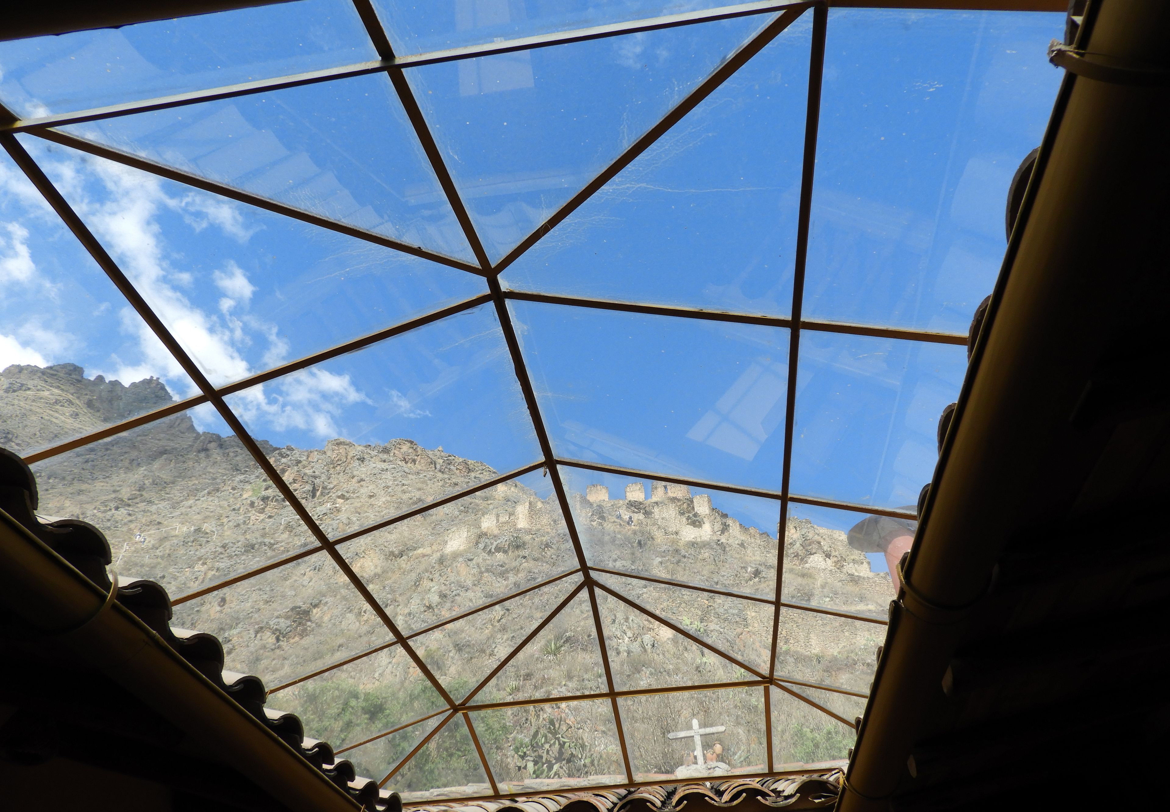 Uitzicht door een geometrisch raamontwerp, waarop een bergachtig landschap met historische ruïnes te zien is.