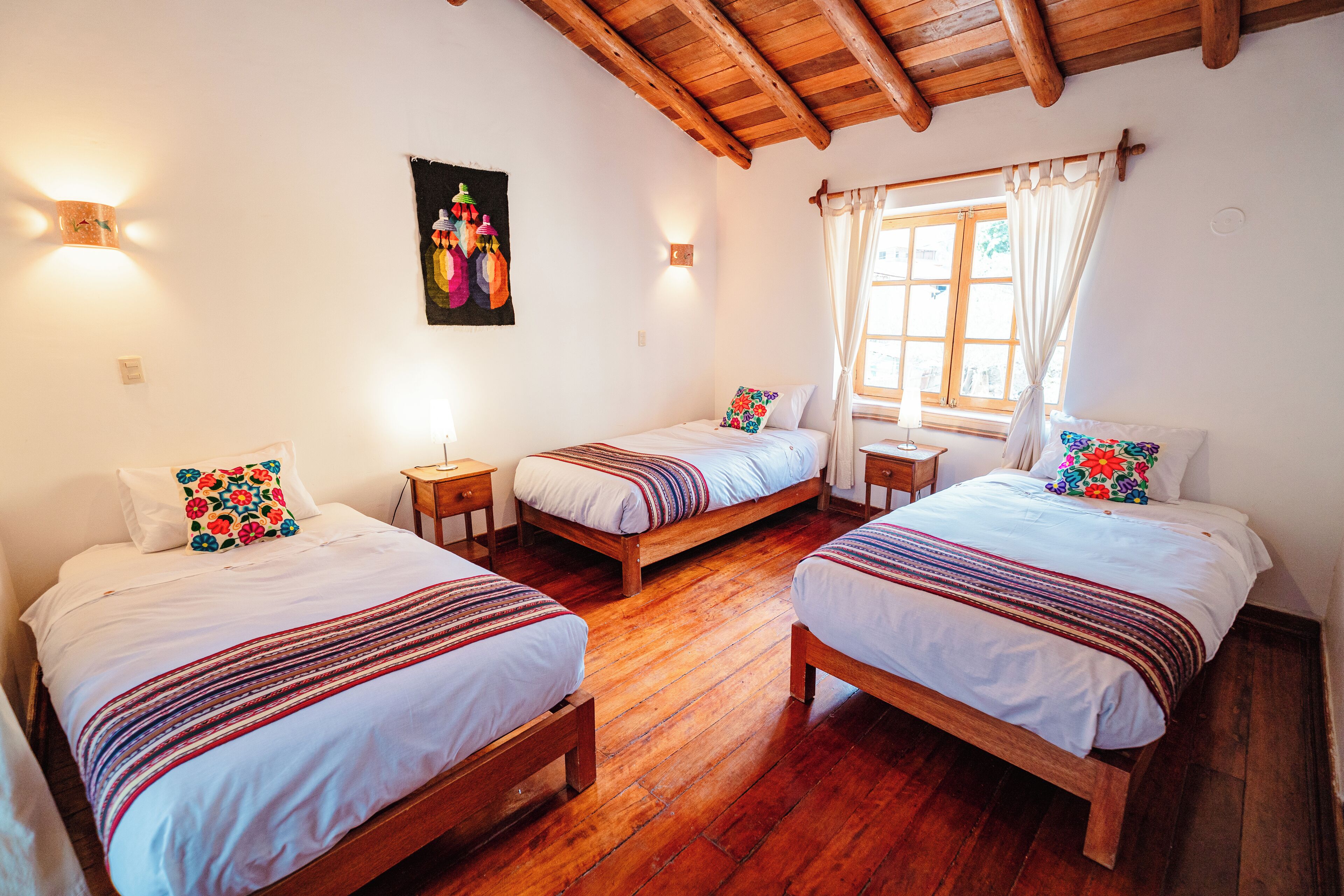 Una habitación con tres camas de madera, cada una con un edredón blanco y almohadas de colores. Tiene techo de madera y una ventana con cortinas.