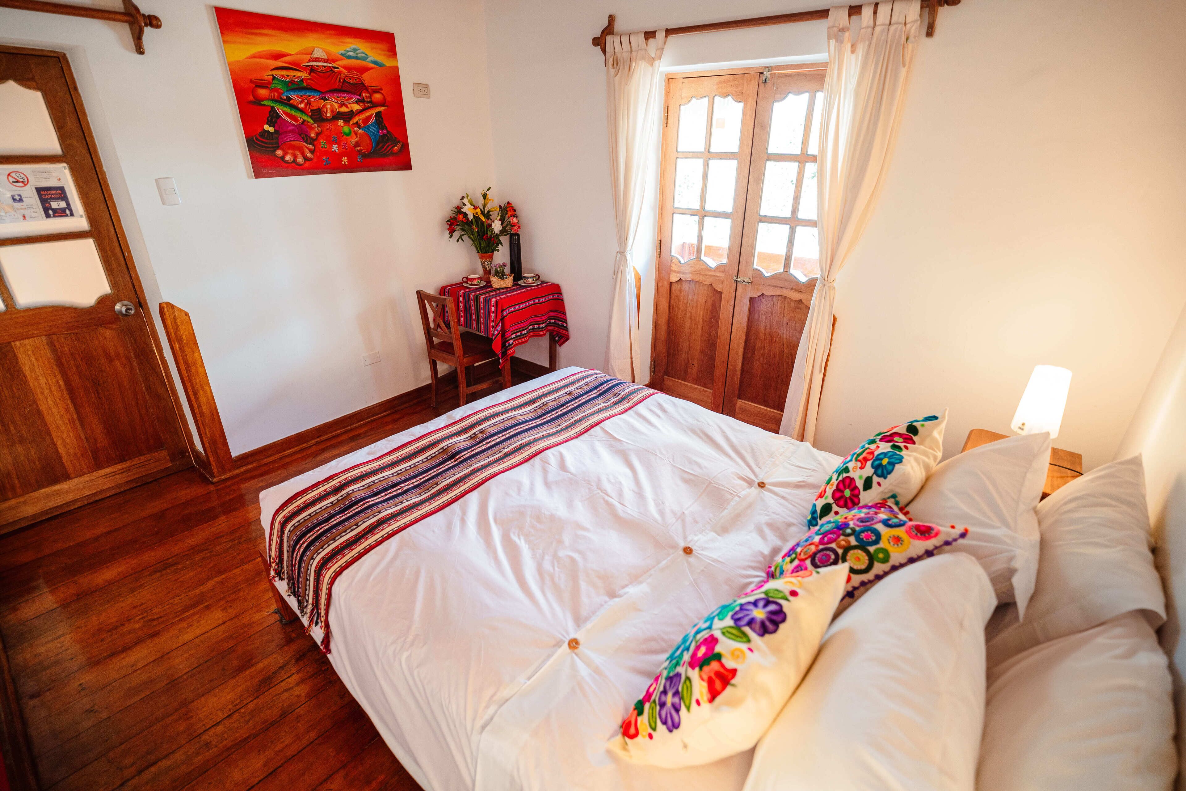 A bedroom with a large bed, wooden floor, a desk with a chair, a window with curtains, and a painting on the wall.