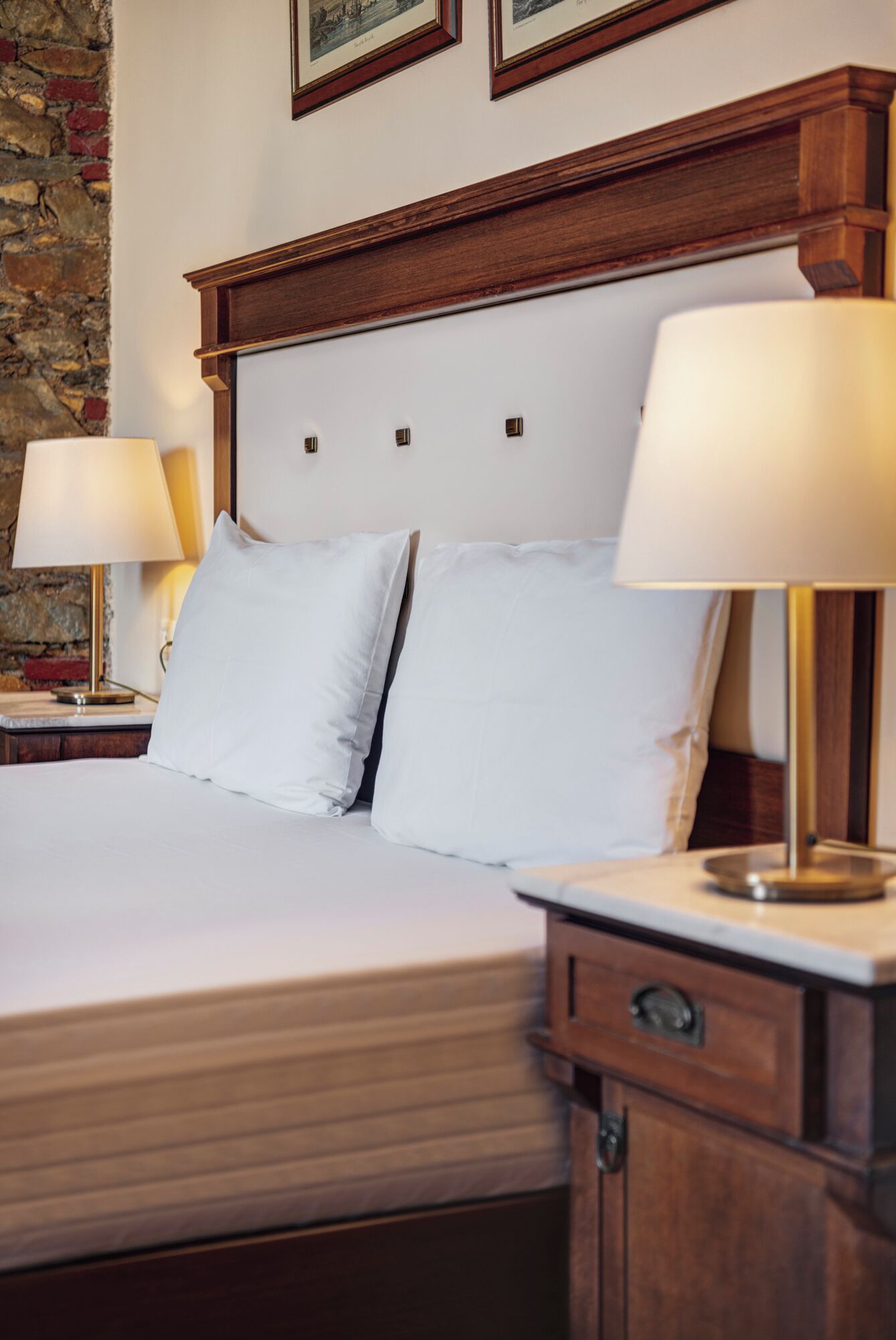 Una cama bien hecha con sábanas blancas, cabecera de madera, dos lámparas de noche y una mesita de noche con lámpara.