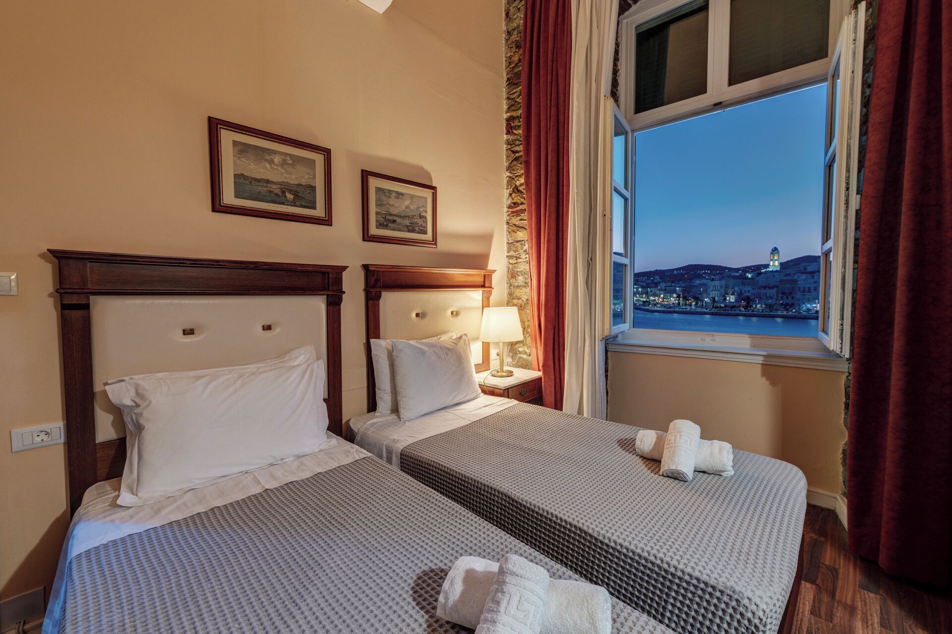 Quarto de hotel com duas camas, uma mesa de cabeceira com abajur, uma janela com vista para a cidade e quadros nas paredes.