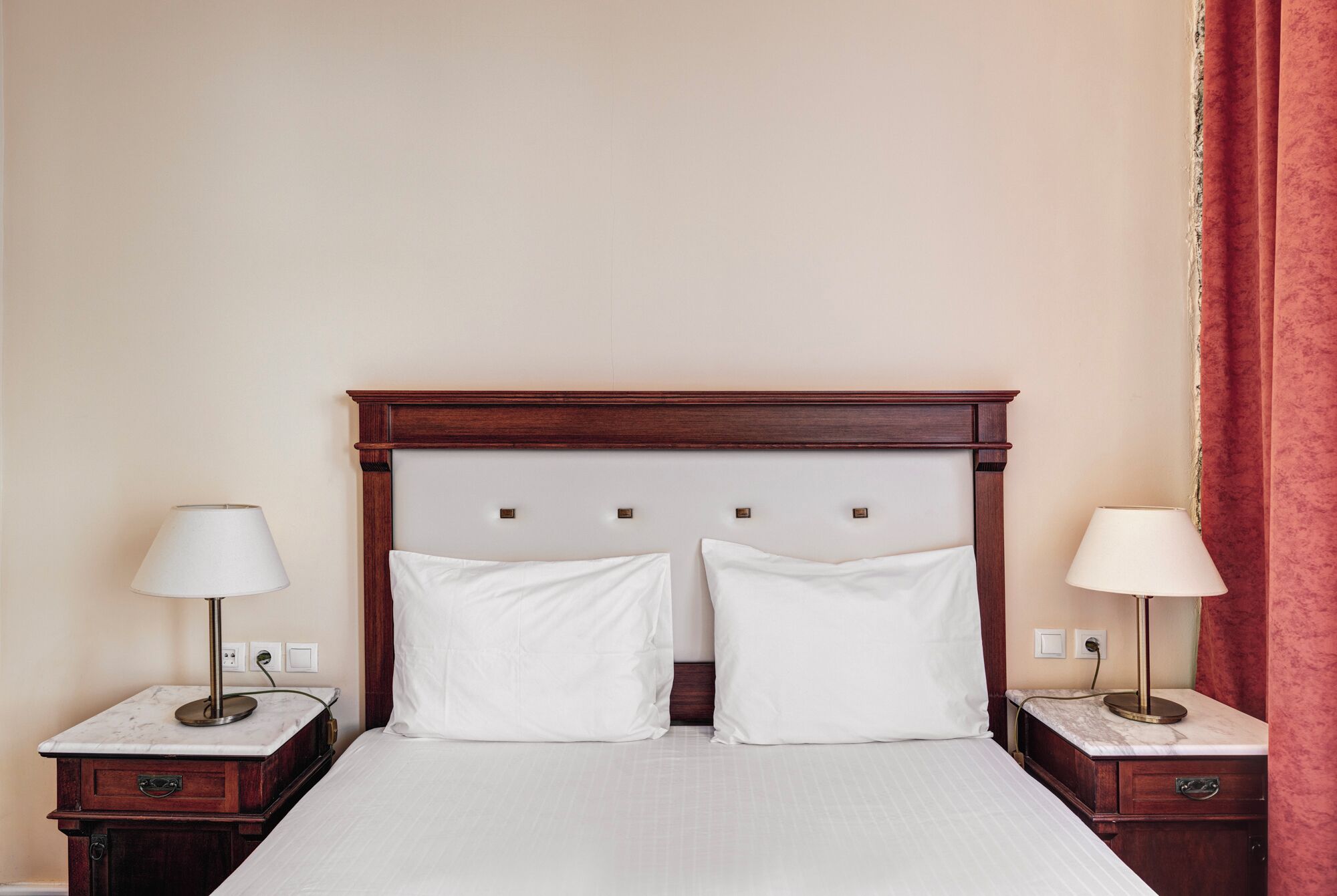 A hotel room with a wooden headboard, two bedside tables with lamps, a bed with white bedding, and a red curtain.