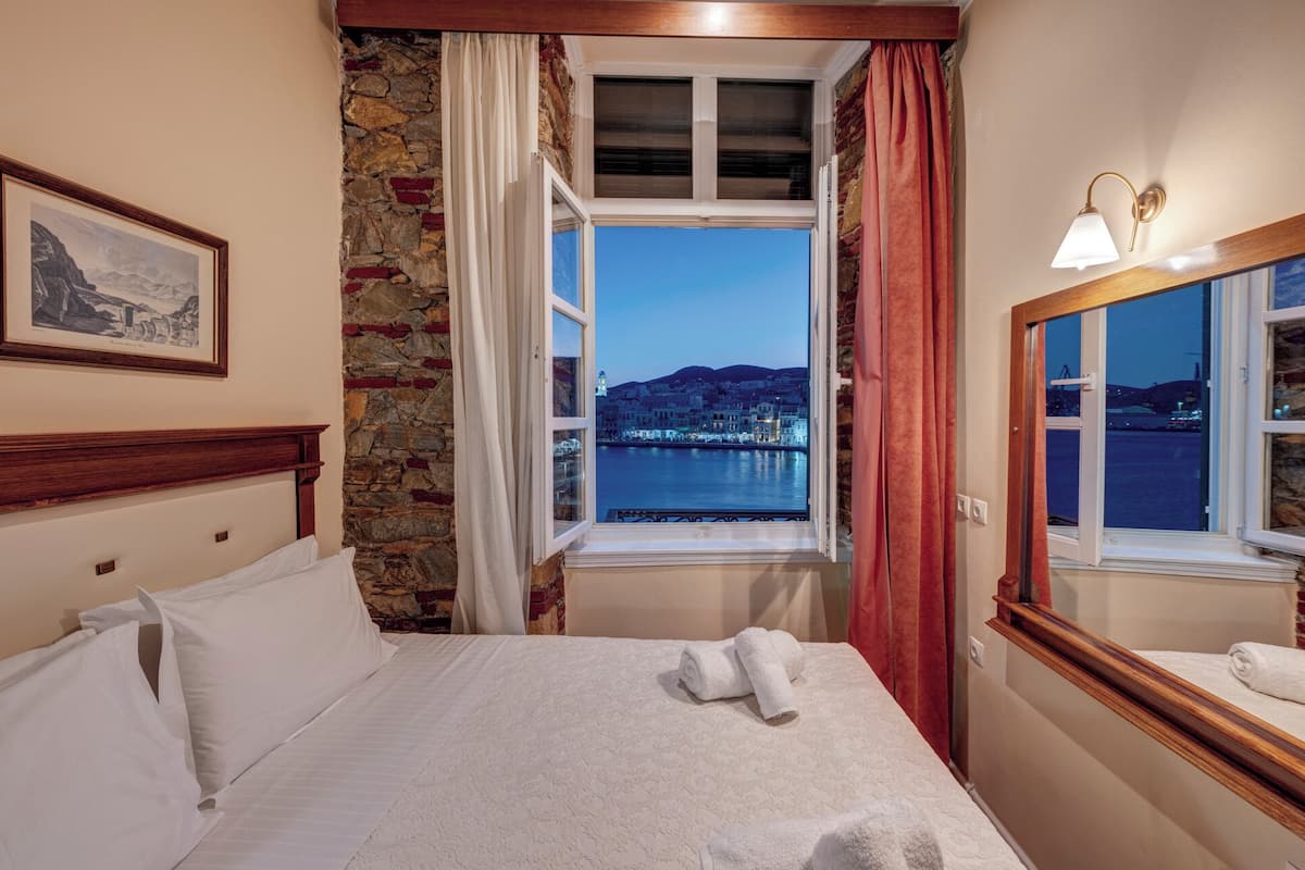 A hotel room with a bed, a window with a view of a cityscape and water, a painting on the wall, and a lamp.