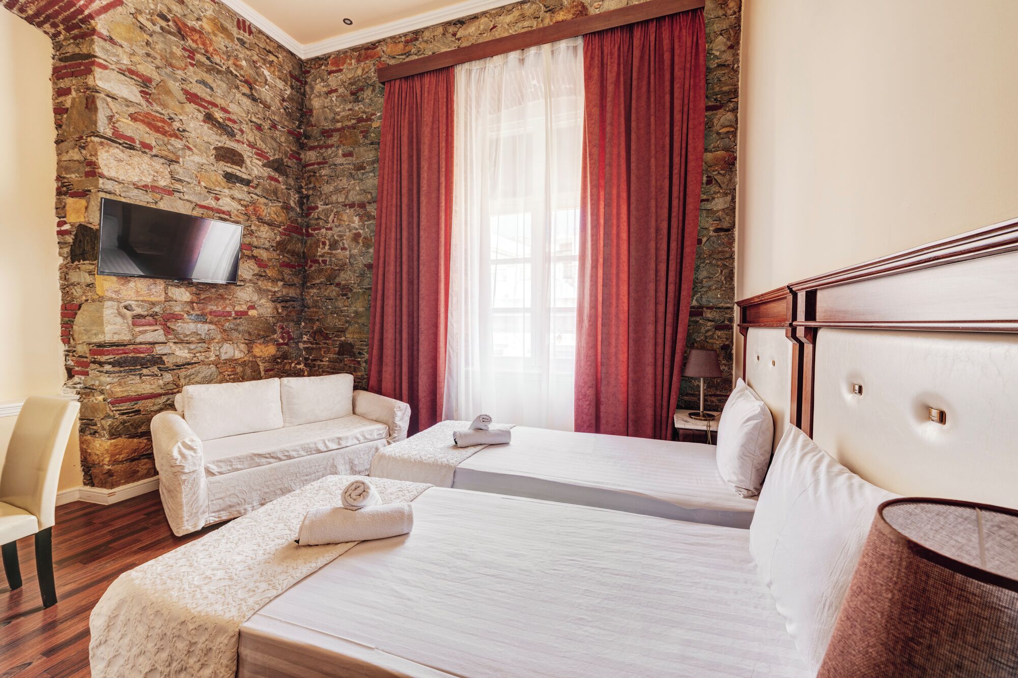 Habitación de hotel con pared de piedra, televisor montado en la pared, cama con cabecera blanca, sofá y ventana con cortinas rojas.