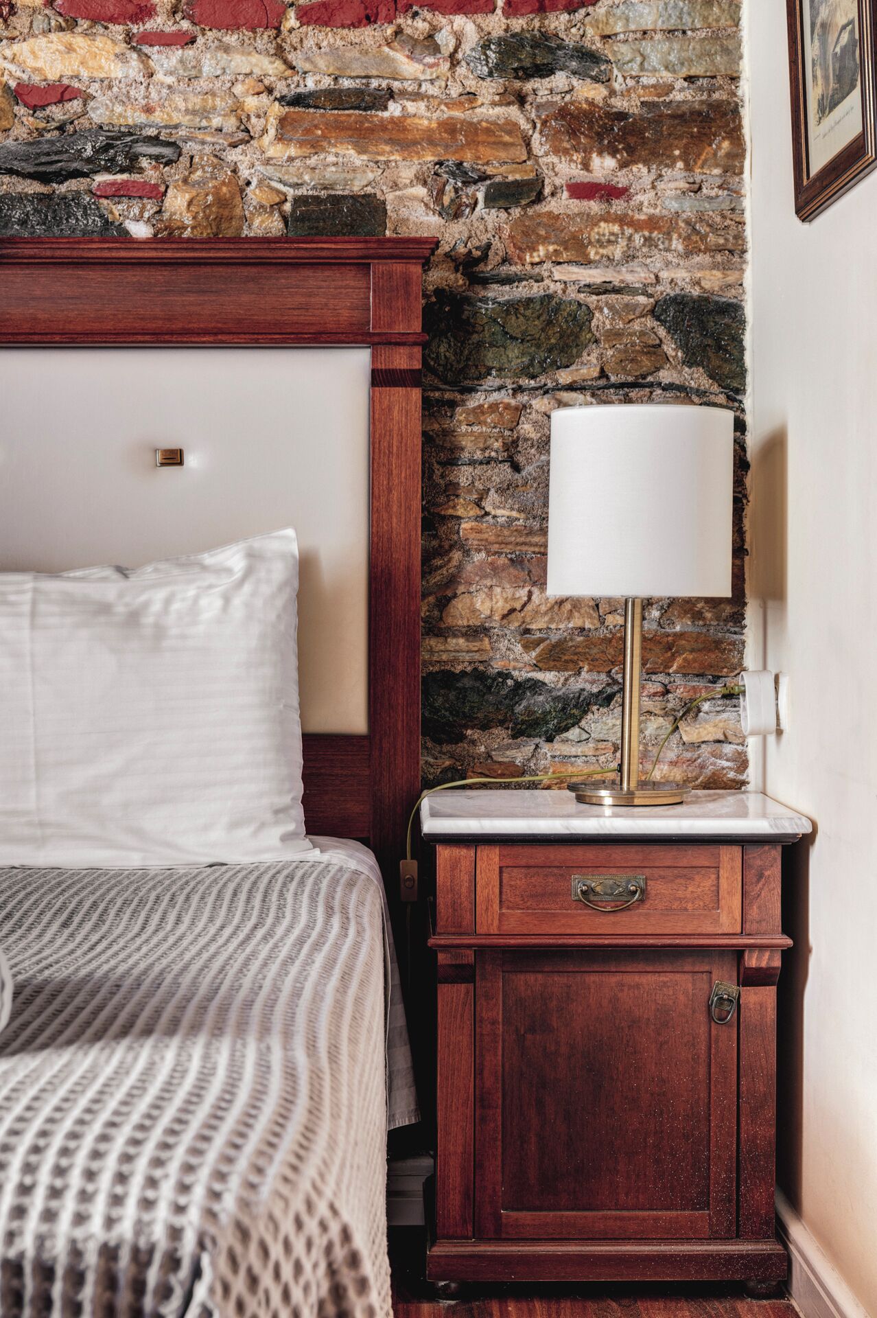 A bedroom with a stone wall, a wooden nightstand, a bed with white linens, and a white lamp.