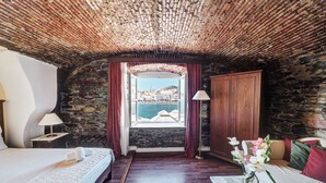Une pièce avec un plafond en pierre, un lit, une armoire en bois, une fenêtre donnant sur la mer et un vase rempli de fleurs.