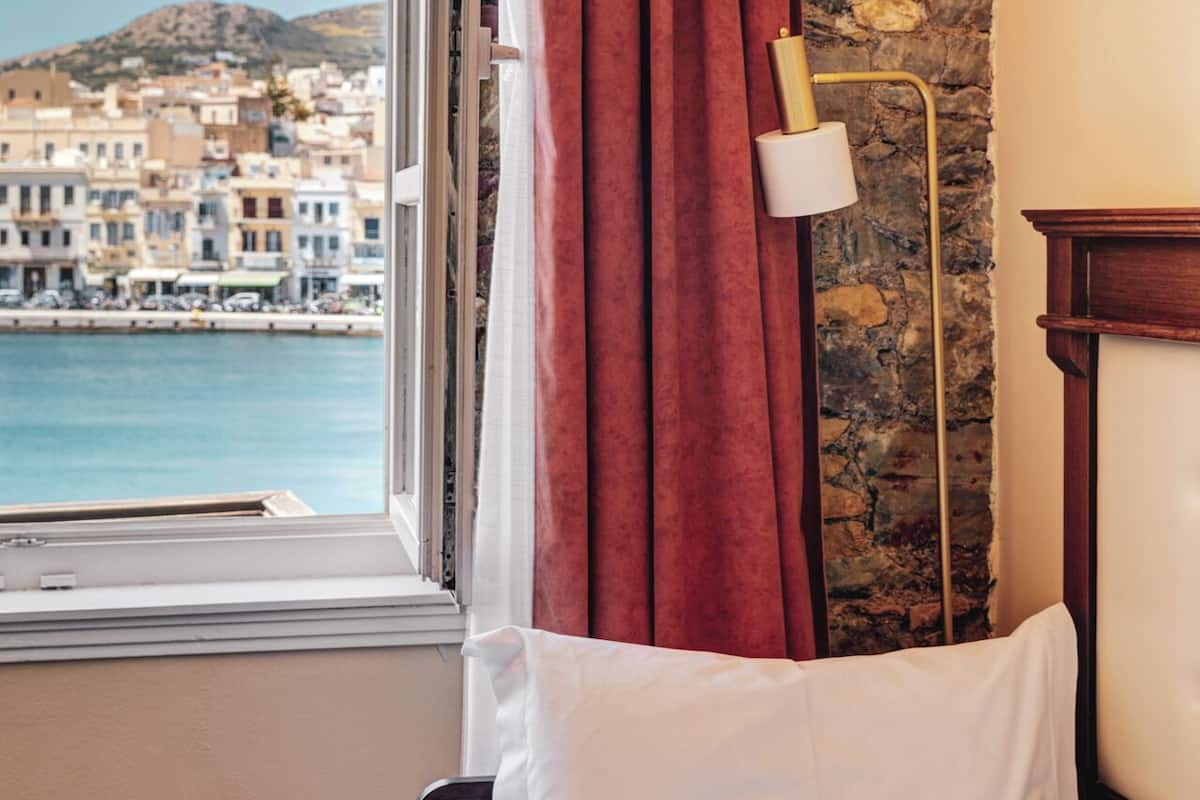 A room with a bed, a window with red curtains, a lamp, and a view of a coastal town.
