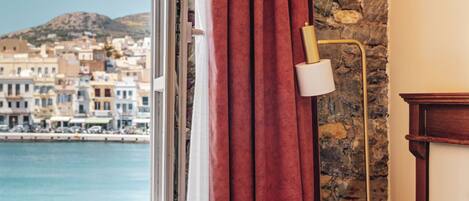 A room with a bed, a window with red curtains, a lamp, and a view of a coastal town.