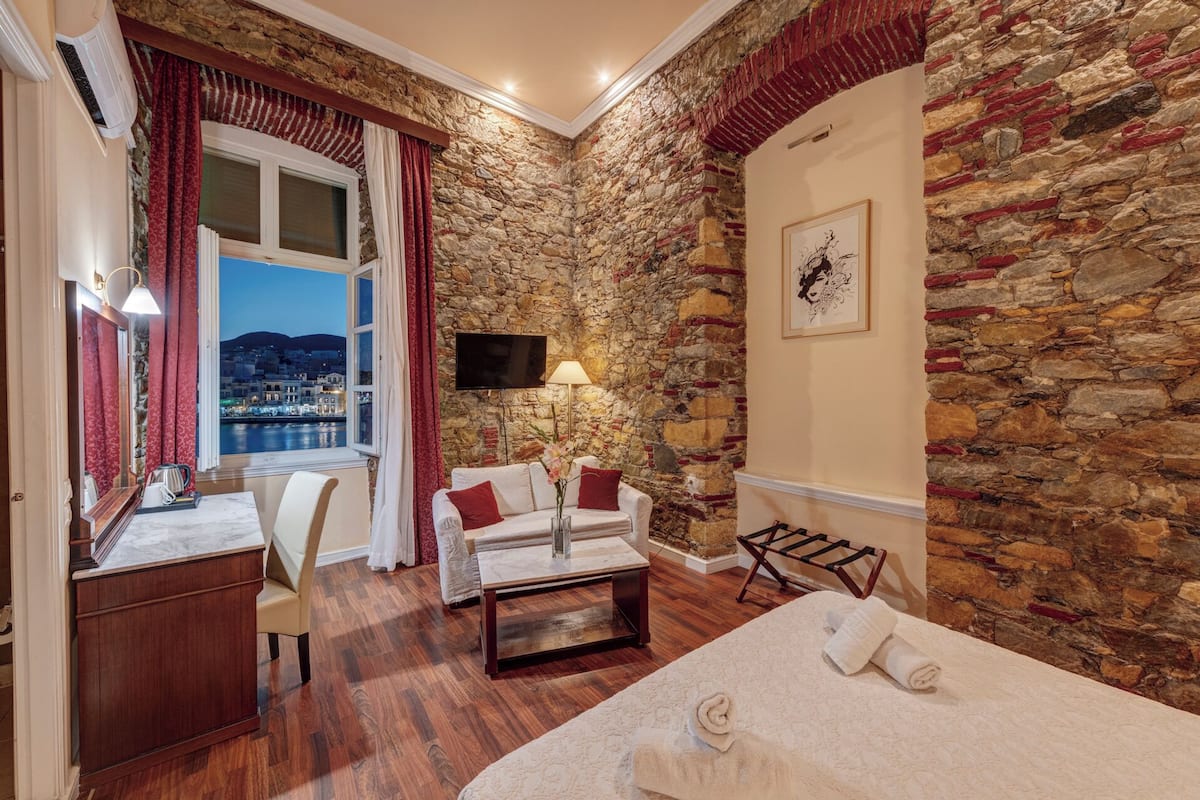 A hotel room with a stone wall, a bed, a desk, a chair, a TV, and a view of a cityscape.