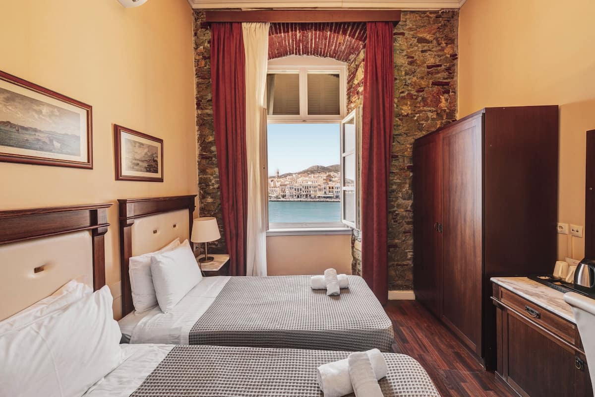 A hotel room with a bed, a nightstand, a wardrobe, and a window with a view of a coastal town.