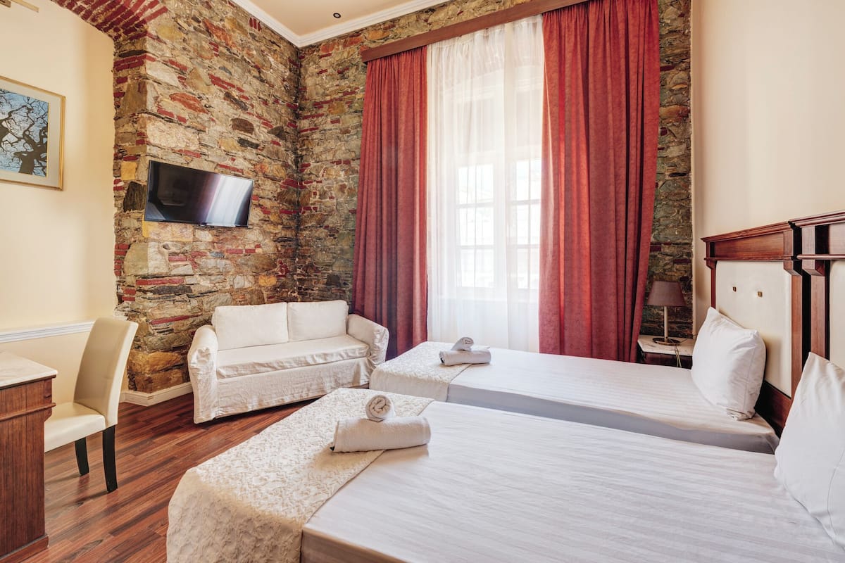 A hotel room with a stone wall, two beds, a TV, a desk, and a chair.