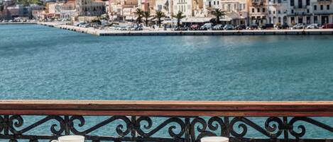 Una terraza frente al mar con una mesa y sillas, con vista a un pueblo costero y una colina al fondo.