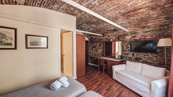 A room with a brick ceiling, a white sofa, a bed with a patterned blanket, a desk with a chair, and a TV mounted on the wall.