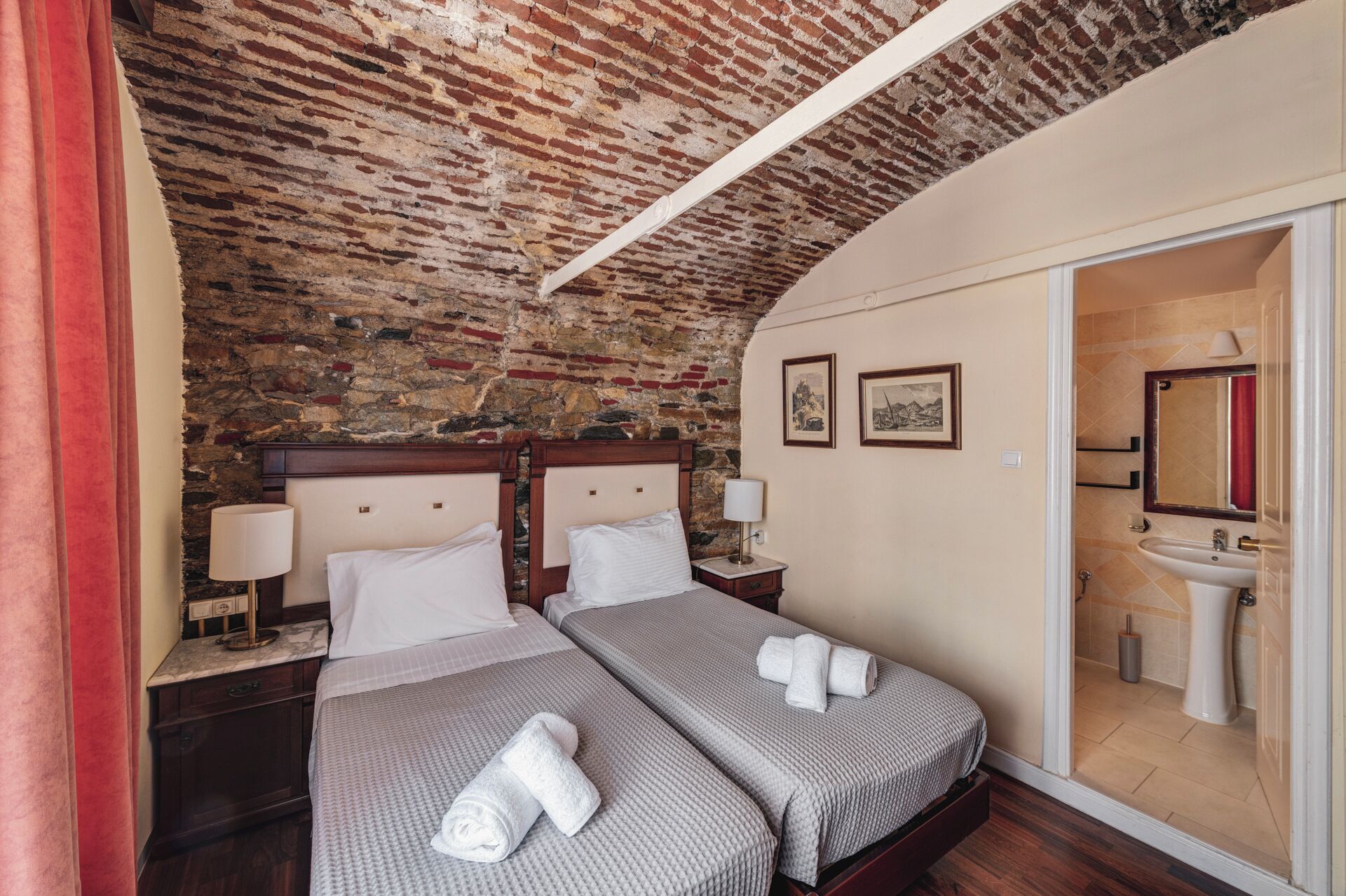 A hotel room with a brick ceiling, two beds with white linens, a nightstand with a lamp, and a bathroom visible through an open door.