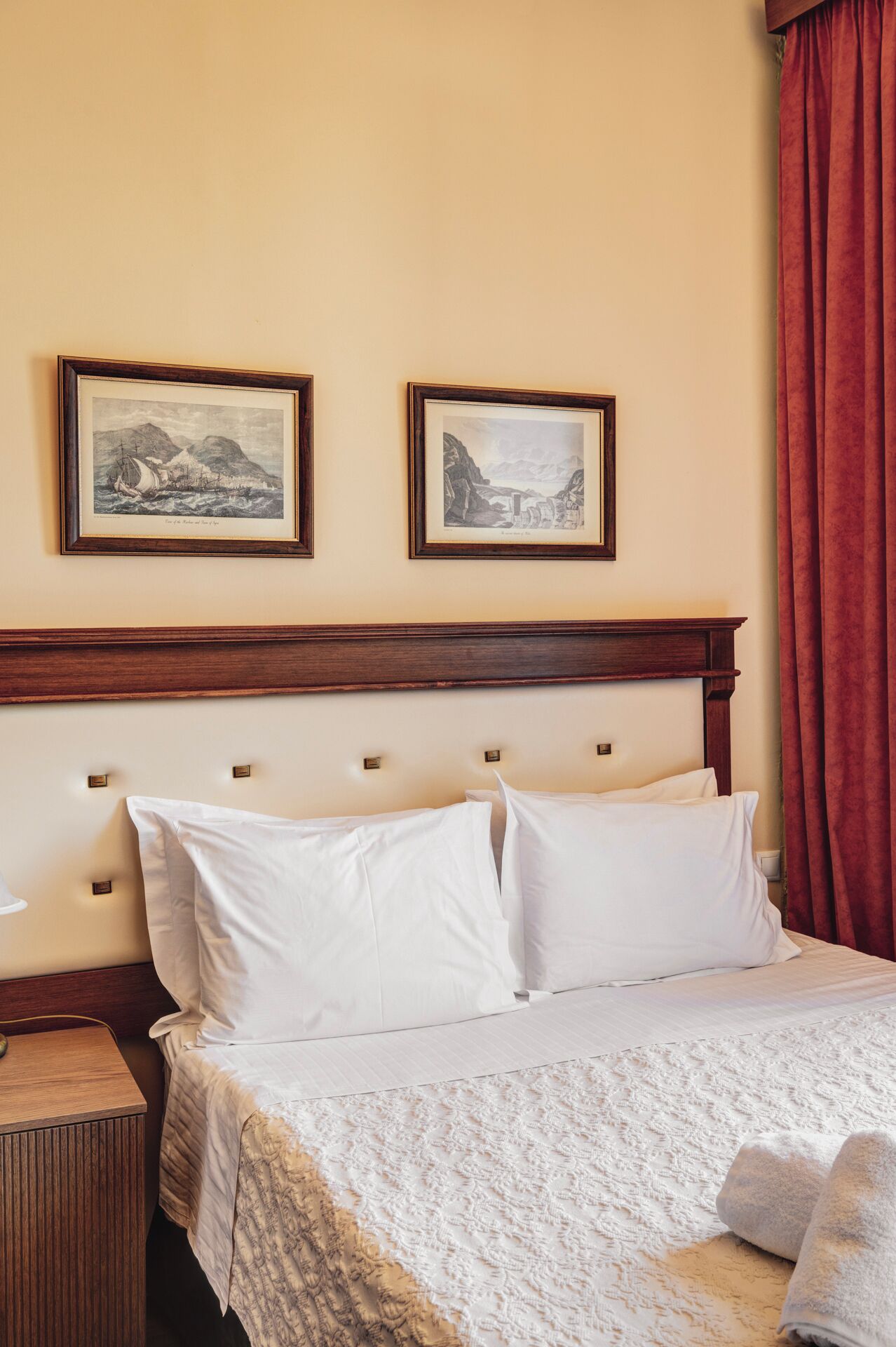 Una cama bien hecha con sábanas blancas y una manta estampada, una mesita de noche de madera con una lámpara y dos cuadros enmarcados sobre la cama.