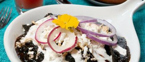 A bowl of enchiladas topped with sour cream and onions, a glass of red drink, and a bread roll on a turquoise tablecloth.