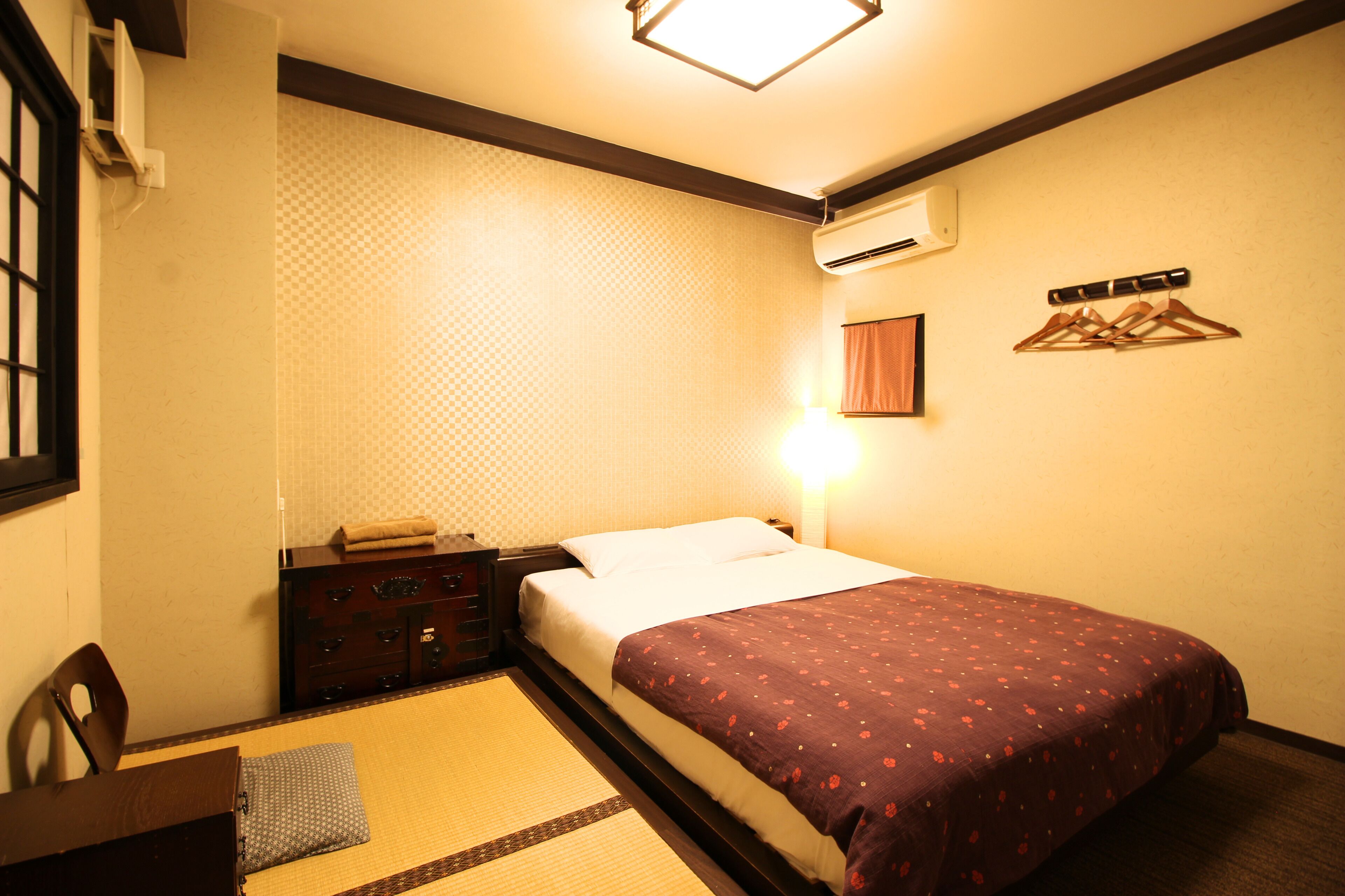 A hotel room with a bed, a bedside table, a chair, a wall-mounted air conditioner, and a wall-mounted light fixture.