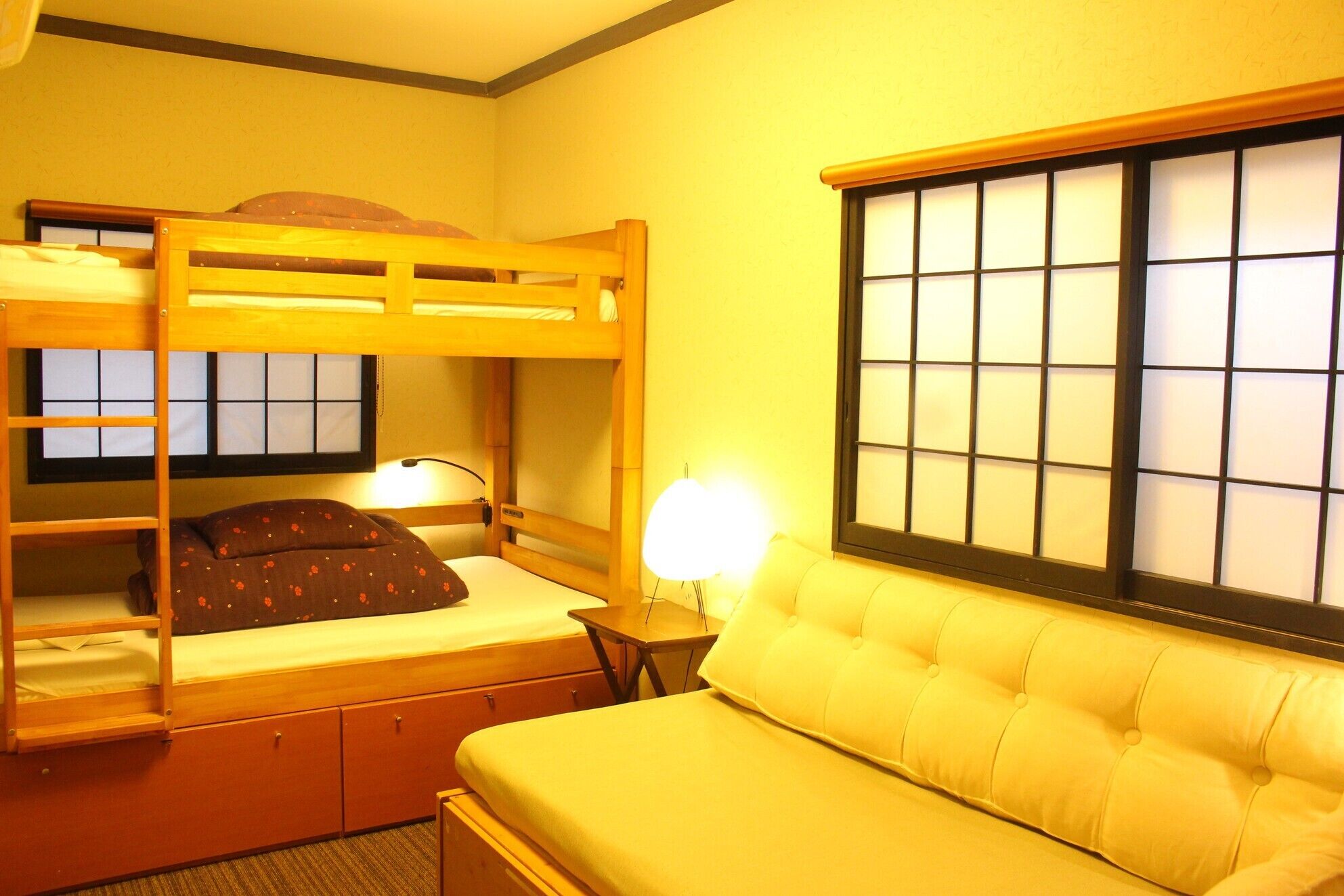 A room with bunk beds, a sofa, a bedside table with a lamp, and a window with a grid pattern.