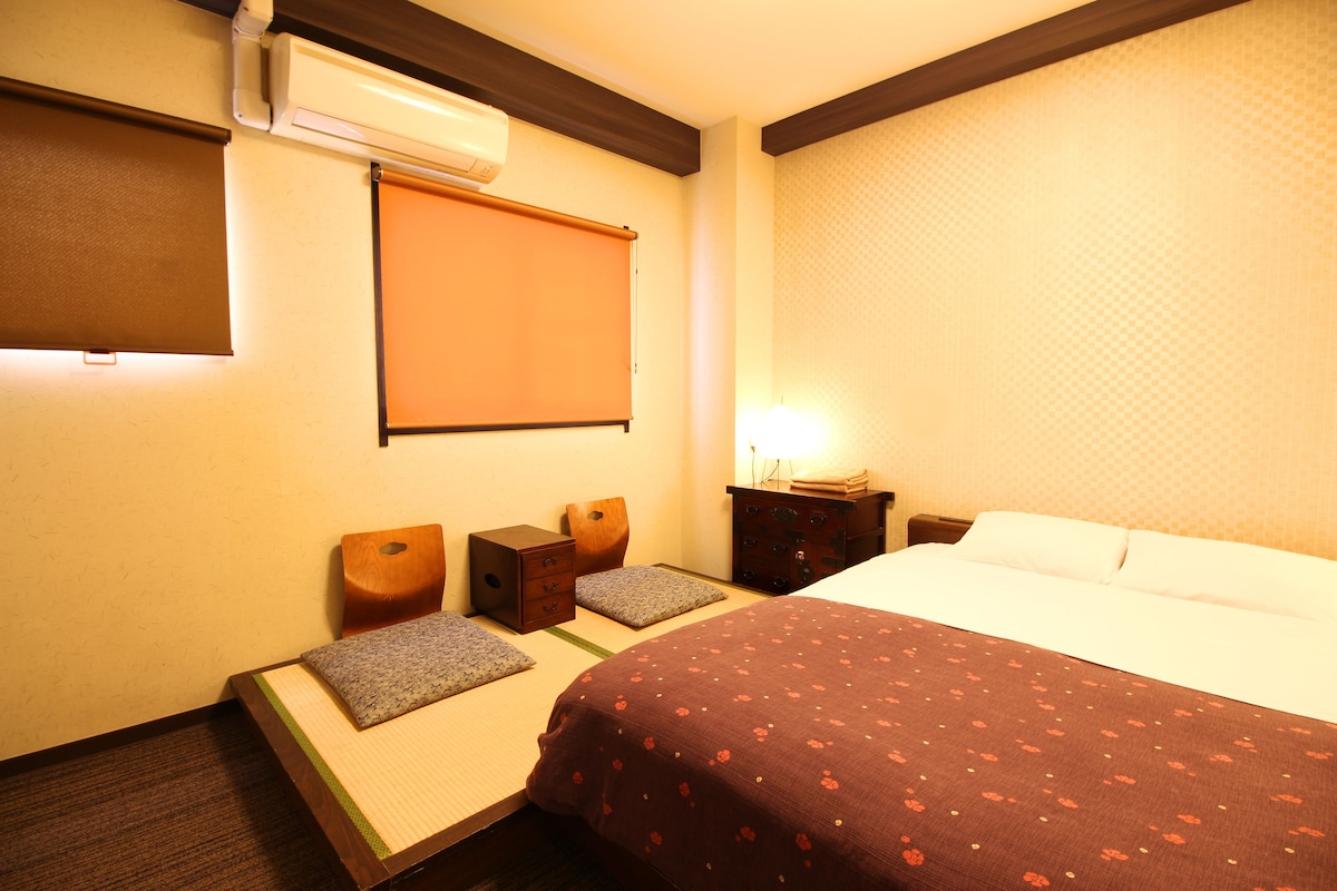 A hotel room with a bed, a small table, a chair, a bedside table, and a wall-mounted air conditioner.