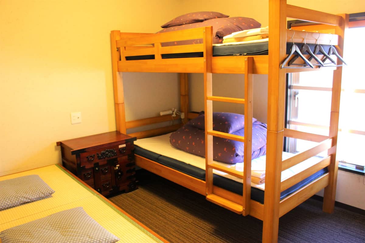 Una cama litera con estructura de madera, un pequeño aparador de madera y una ventana con vistas.