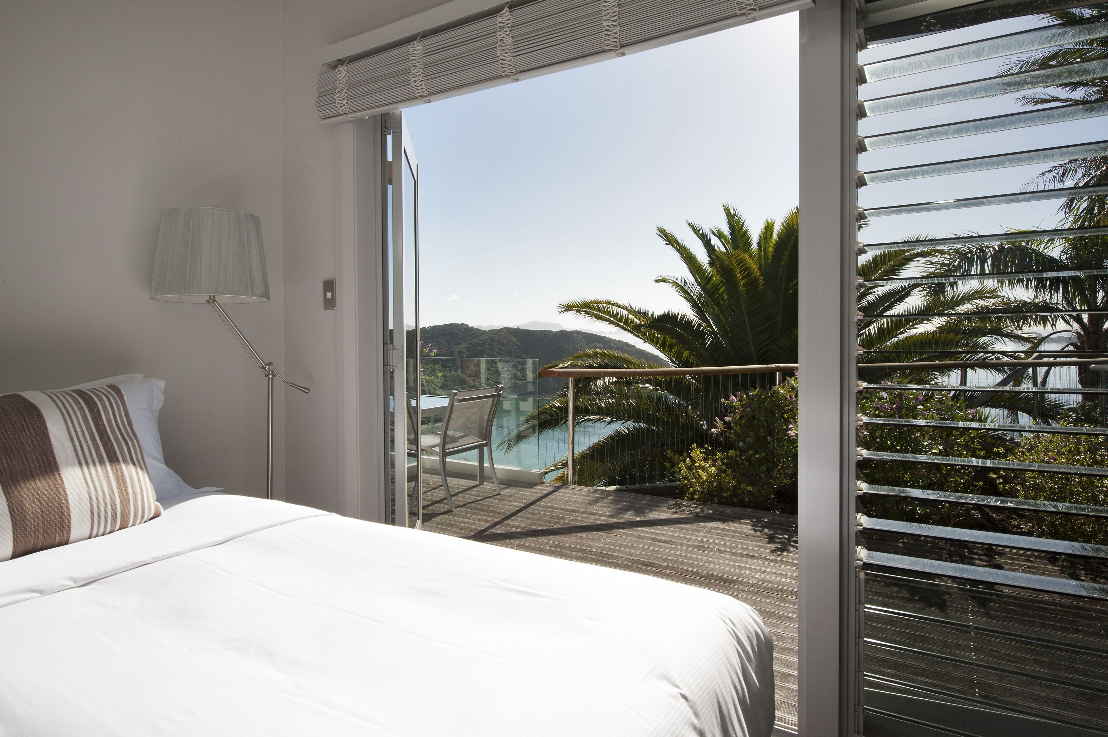 A bedroom with a large bed, a nightstand, a lamp, and a sliding glass door leading to a balcony with a view of palm trees and a body of water.