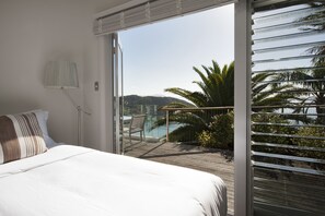 A bedroom with a large bed, a nightstand, a lamp, and a sliding glass door leading to a balcony with a view of palm trees and a body of water. - Cloud 9 Luxury Villa (Russell)