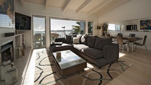 A spacious living area with a large sofa, a coffee table, and a dining table set for four. A large window offers a view of the outdoors. - Cloud 9 Luxury Villa (Russell)