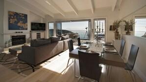 A modern living room with a sofa, dining table, and chairs. There is a fireplace, a television, and a large window with a view of the sea. - Cloud 9 Luxury Villa (Russell)