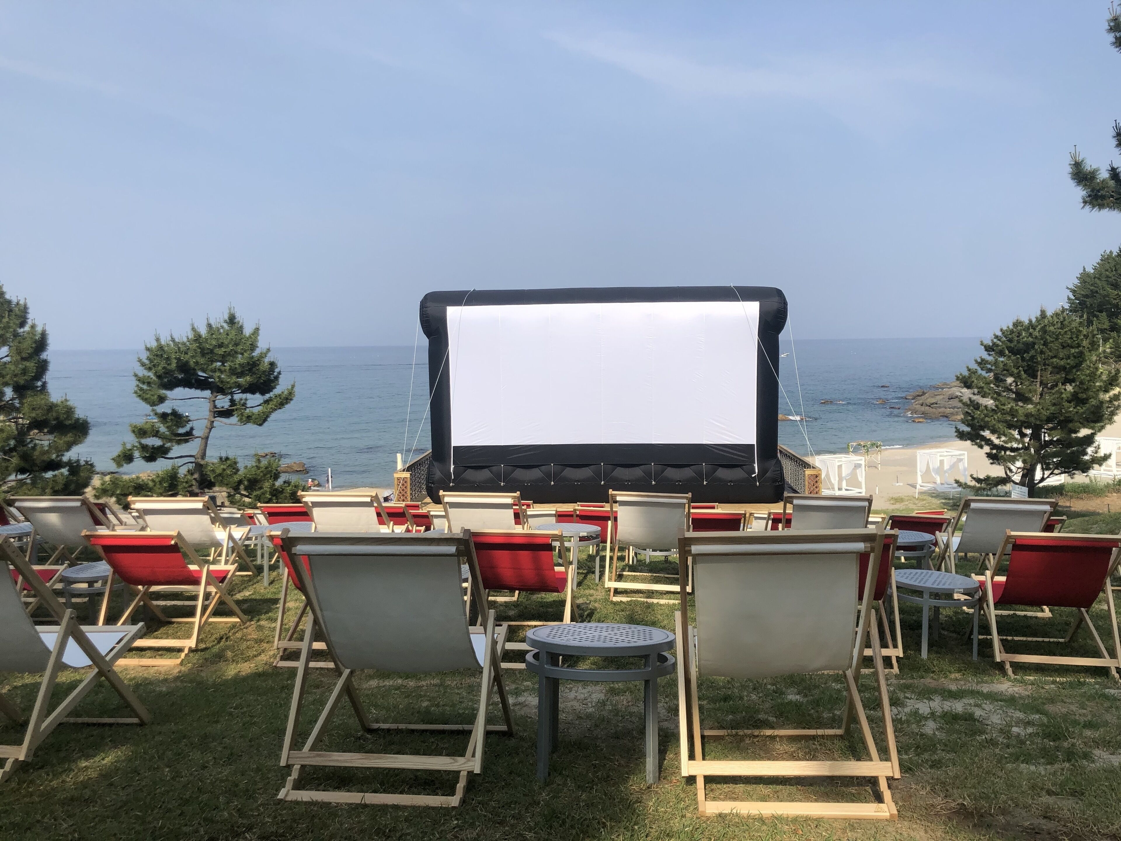 Un espace extérieur aménagé avec des sièges, un grand écran, des chaises rouges et grises, et une vue sur la mer.