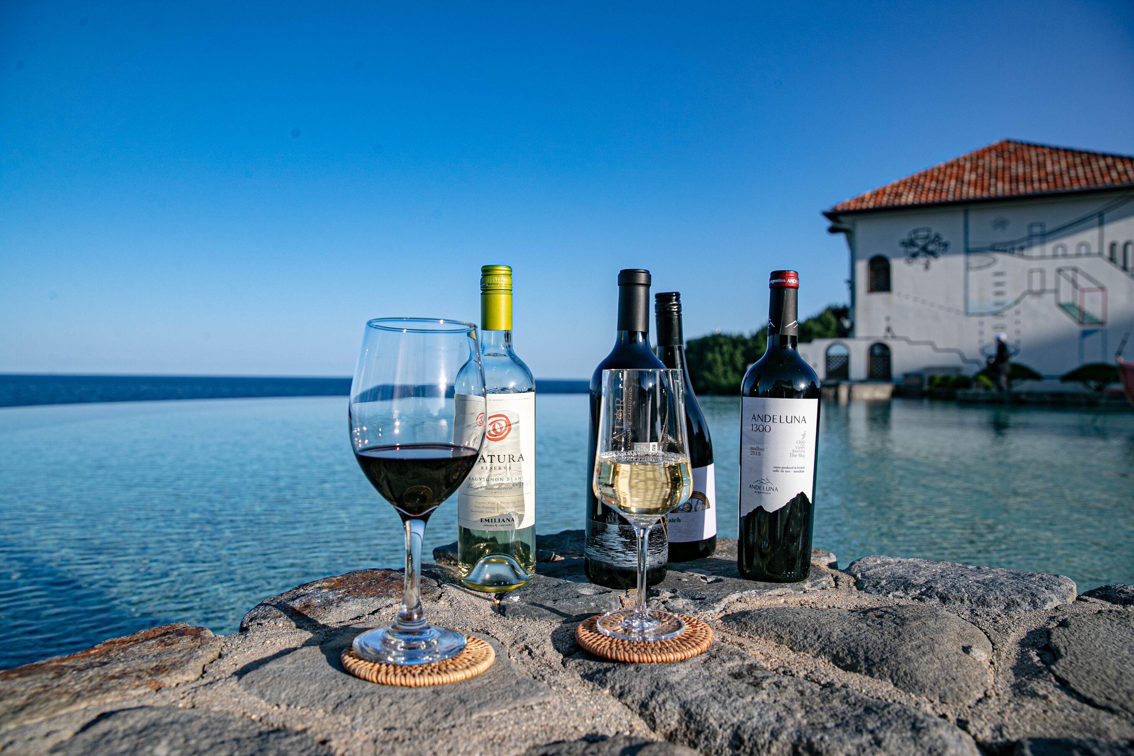 A stone table with four wine bottles and two glasses, overlooking a body of water and a building with a tiled roof.