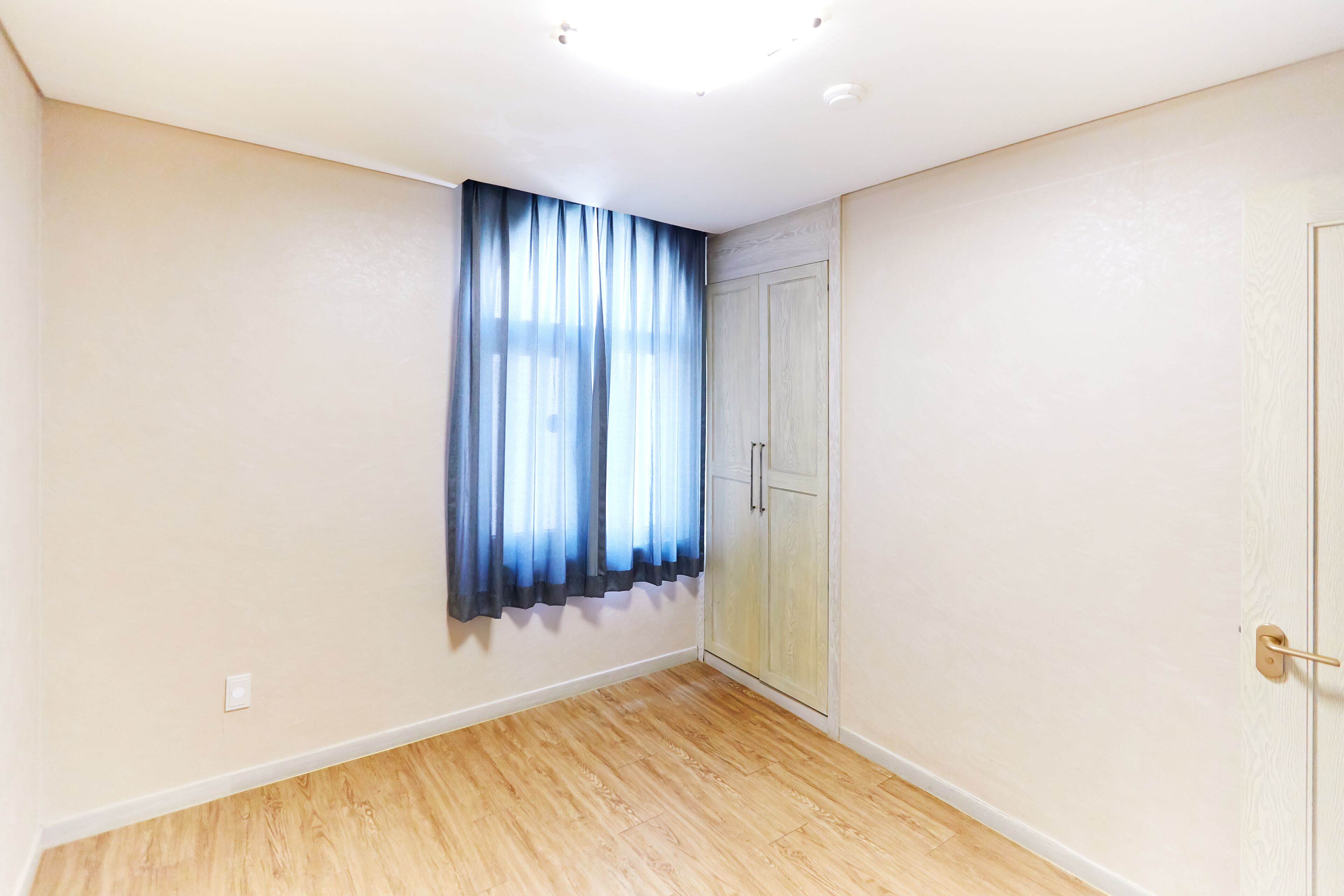 A room with a wooden floor, a door, and a window with blue curtains.